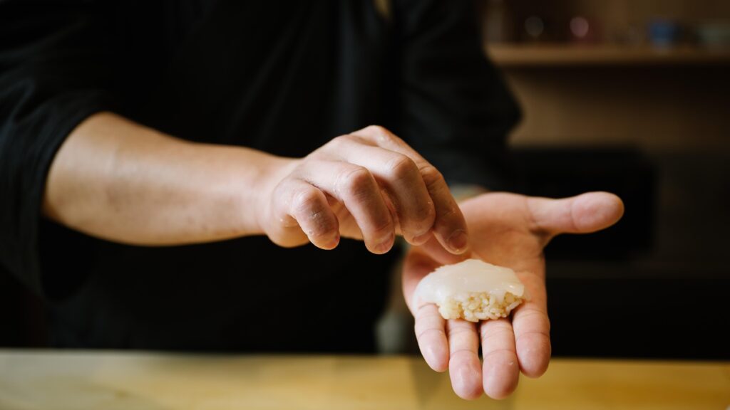 Sushi Masa’s hands carefully assemble a piece of nigiri, placing a delicate slice of pale fish atop a small bed of seasoned rice.