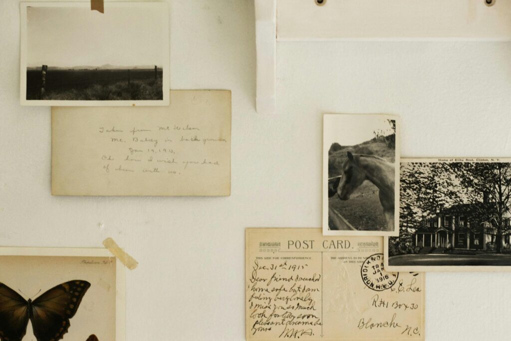 This image captures a nostalgic collection of vintage photographs, handwritten postcards, and a butterfly specimen pinned to a white wall. The aging paper and black-and-white imagery evoke a sense of personal history and quiet longing from the early 20th century.