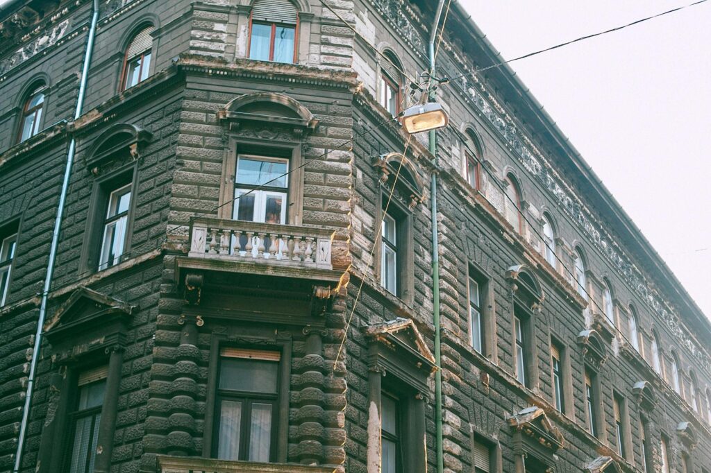 An ornate, multi-story stone building features classic architectural details like pedimented windows and a small stone balcony overlooking a street corner.