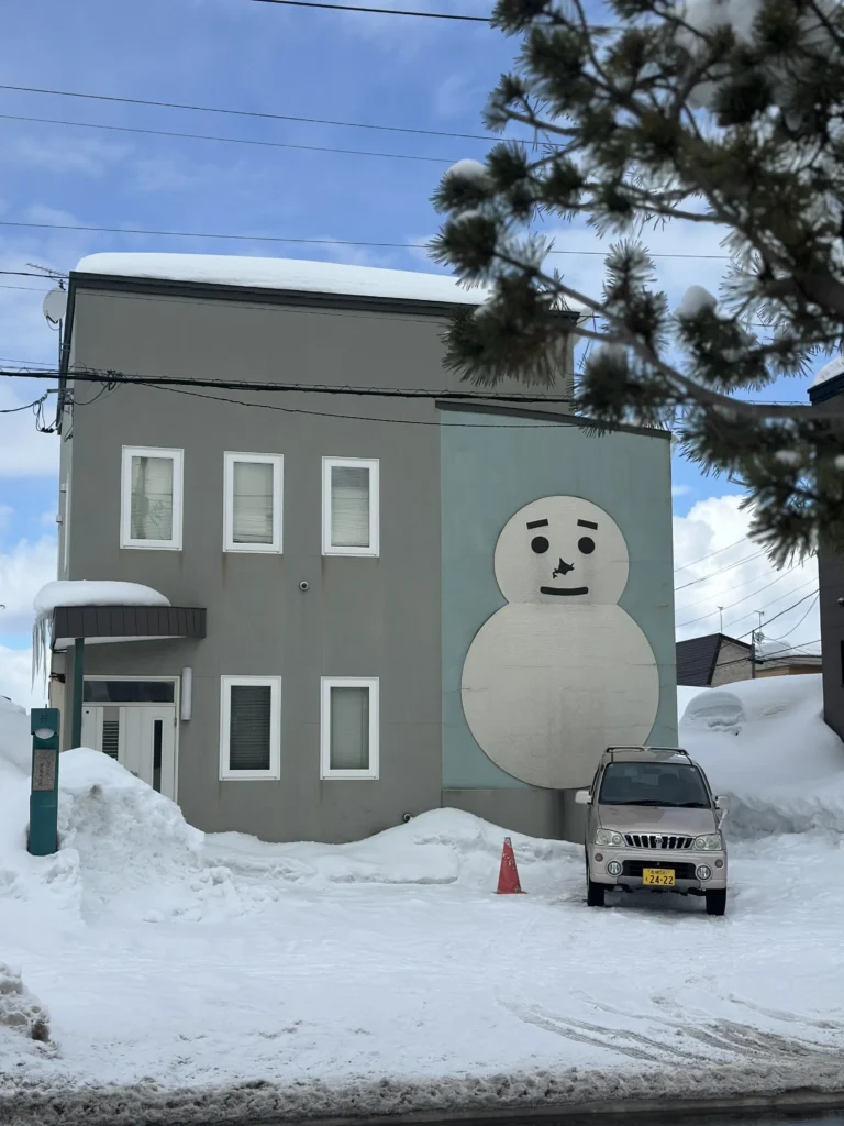 A gray building features a large, minimalist snowman mural on its side, standing prominently next to a gold car parked in a snowy lot. Thick layers of snow cover the ground and the building's roof, emphasizing the cold, wintry atmosphere of the neighborhood.