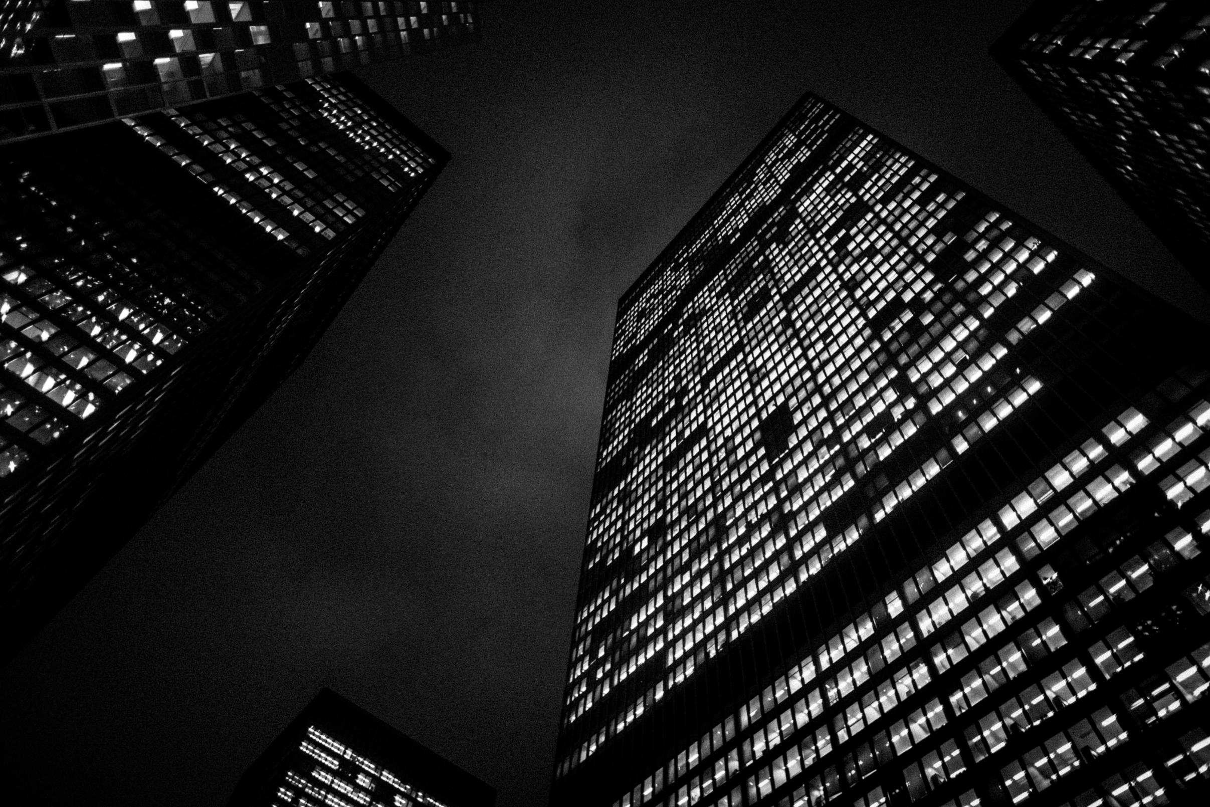 A low-angle, black-and-white shot captures several towering skyscrapers reaching toward a dark, overcast sky. The glowing windows of the buildings create a stark, rhythmic pattern of light against the deep shadows of the urban landscape.