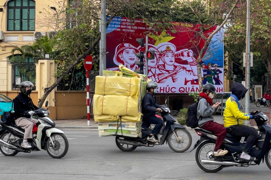 Motorcyclists navigate a busy street in Vietnam, including one rider carrying a massive, towering load of yellow packages.