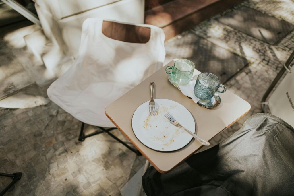 The image shows the remnants of a meal on a small outdoor table, featuring a messy white plate with two forks and two green textured glass mugs.