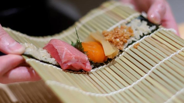 Hands hold a bamboo rolling mat topped with a sheet of nori and seasoned rice. The sushi roll features a variety of fresh ingredients, including a prominent slice of raw tuna, bright orange uni, and crispy toppings.
