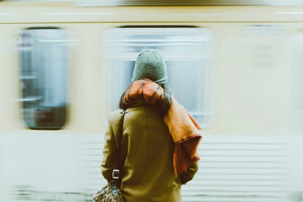 A woman wearing a brown coat and an orange scarf stands still on a platform as a yellow subway train blurs past her. The shot is framed from behind, capturing a quiet moment of stillness.