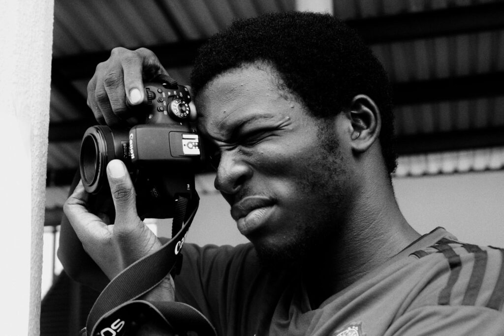 A man is captured in a striking black-and-white close-up as he focuses intensely through the viewfinder of a Canon DSLR camera. His expression is one of deep concentration, with one eye squinted while his hands steady the lens for a shot.