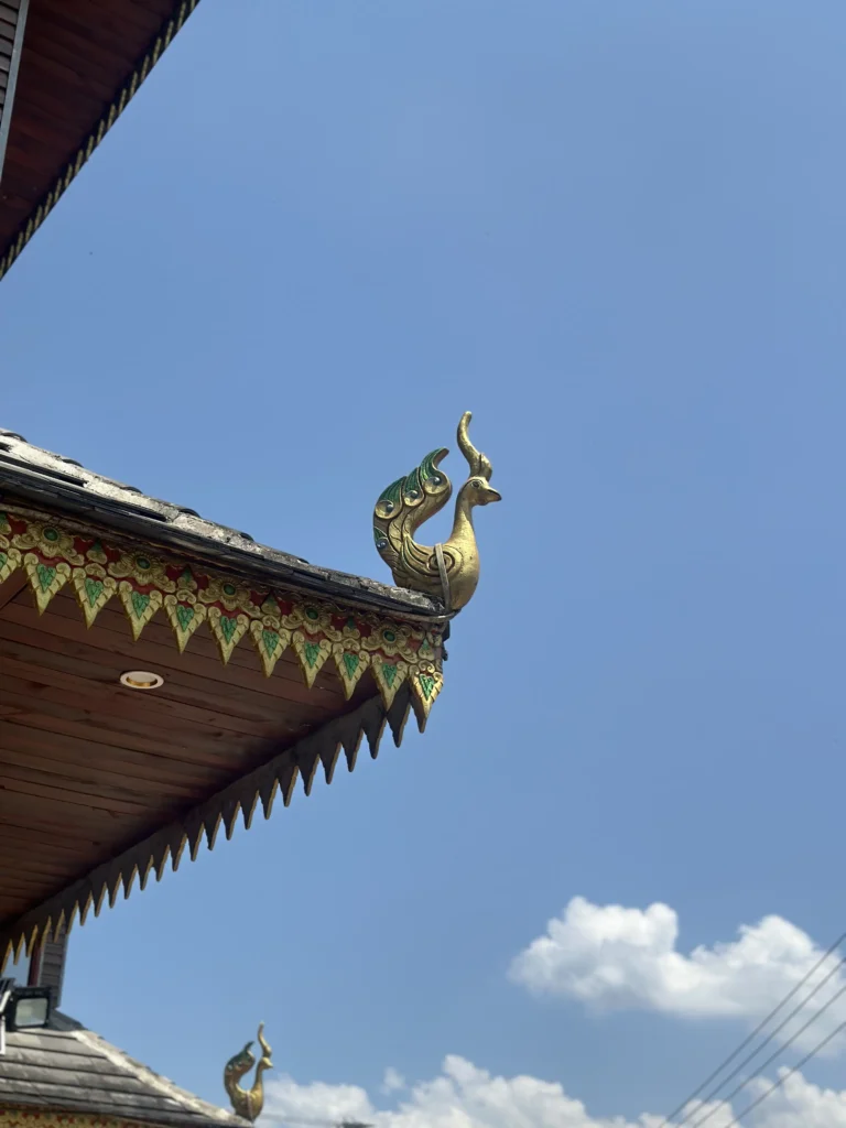 The image shows a gold-colored, bird-like finial, likely a Hamsa or peacock, perched on the corner of a traditional Southeast Asian roof. The roof features intricate, colorful carvings along its edge and is set against a clear, bright blue sky with a few wispy clouds.