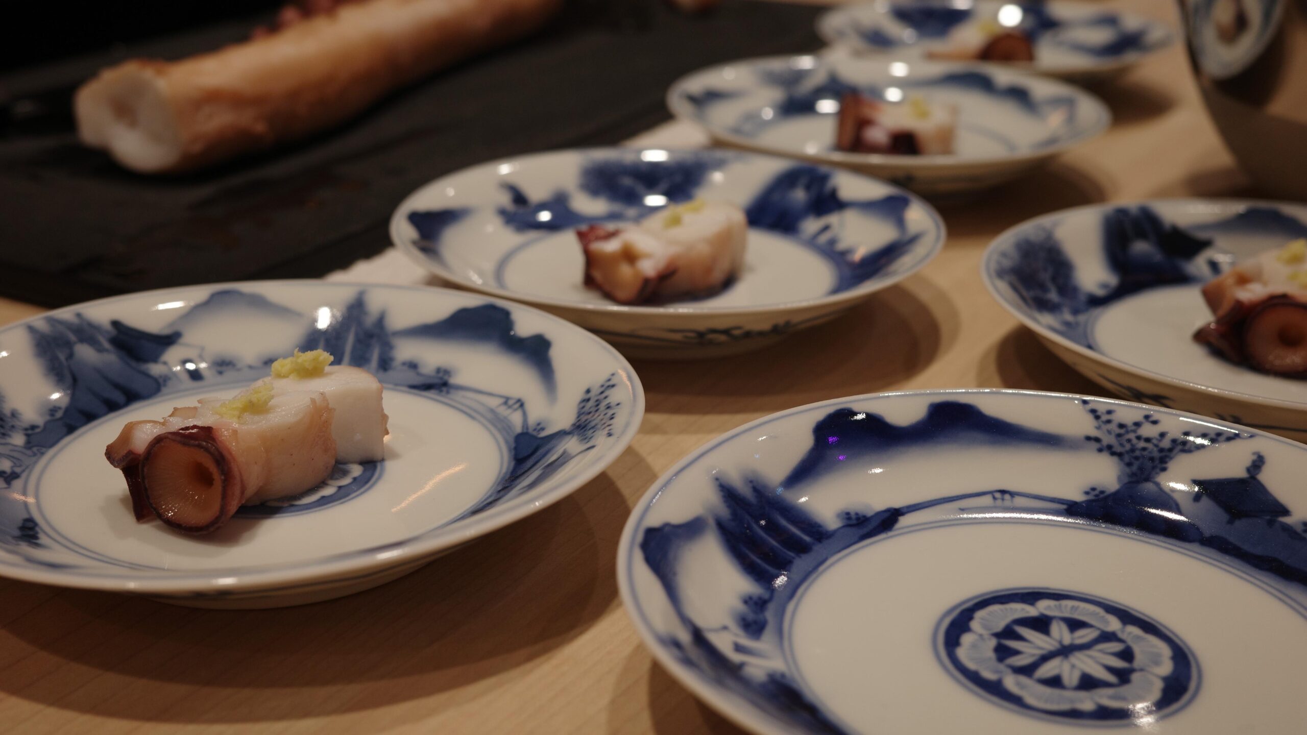 Slices of tender octopus garnished with wasabi are meticulously arranged on elegant blue and white porcelain plates. The shallow depth of field captures several servings prepared on a light wooden counter.
