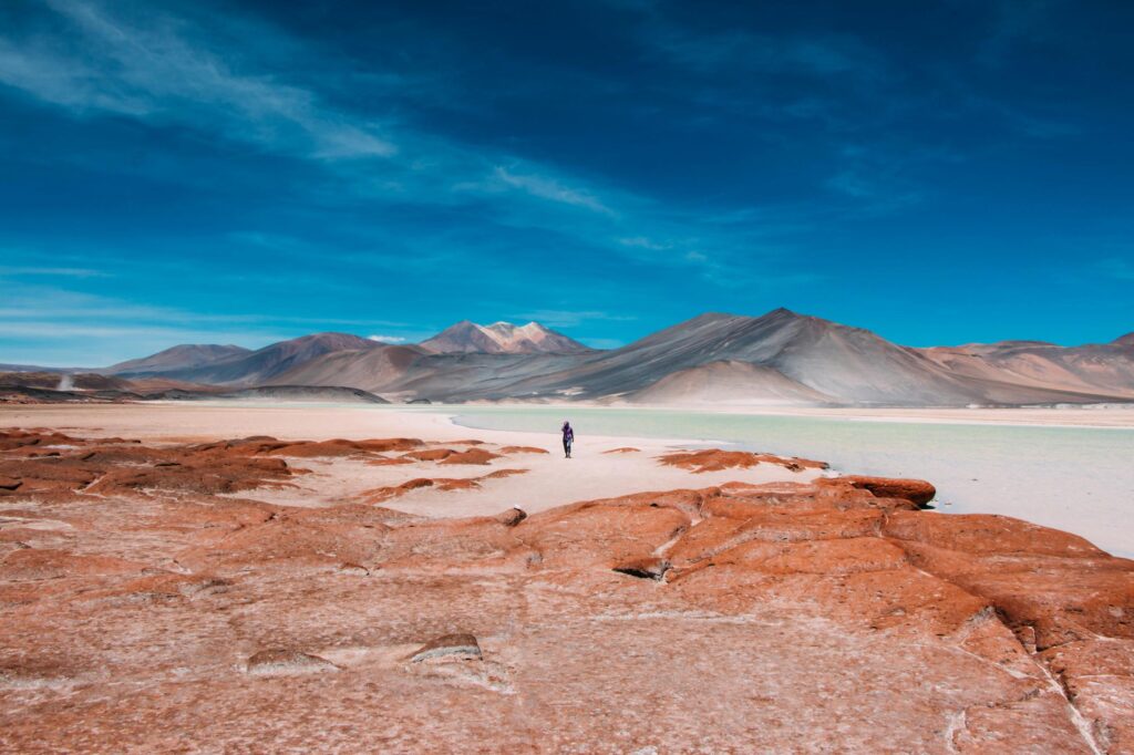 A lone figure stands on a vast, reddish-orange landscape under a bright blue sky streaked with wispy clouds. In the background, rolling grey mountains rise behind a pale, turquoise body of water that cuts through the desert terrain.