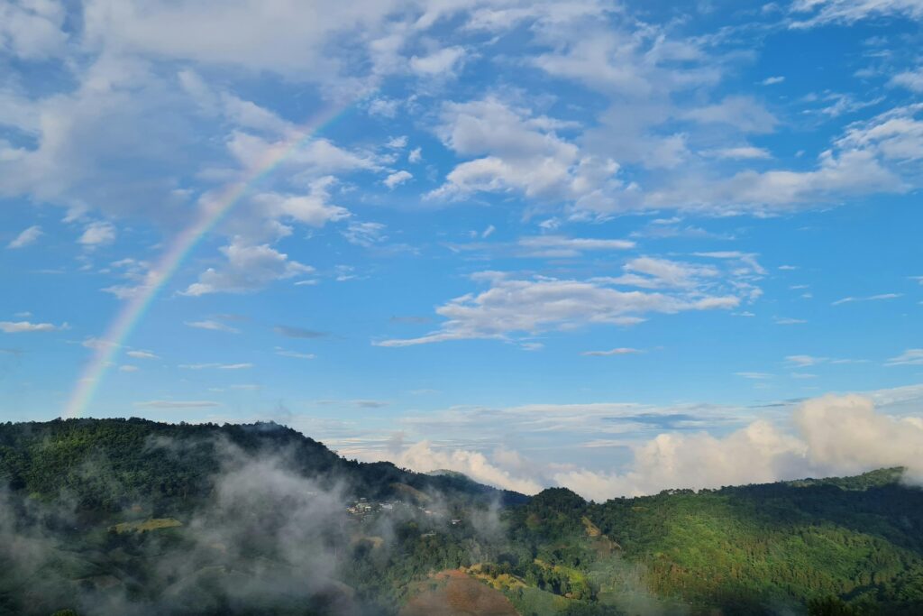 A vibrant rainbow arches across a bright blue sky filled with scattered white clouds. Below, mist and low-lying clouds weave through a series of lush, green forested hills.