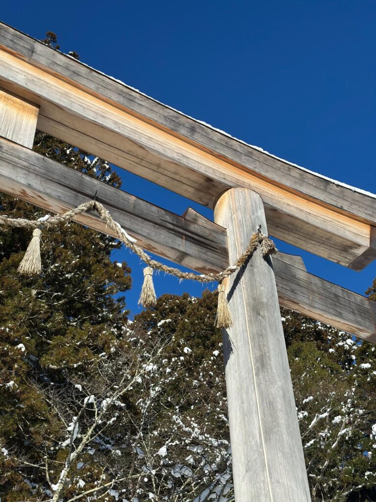 The image shows a close-up, low-angle view of a traditional wooden Shinto torii gate adorned with a straw shimenawa rope. In the background, snow-dusted evergreen trees stand against a crisp, clear blue sky.