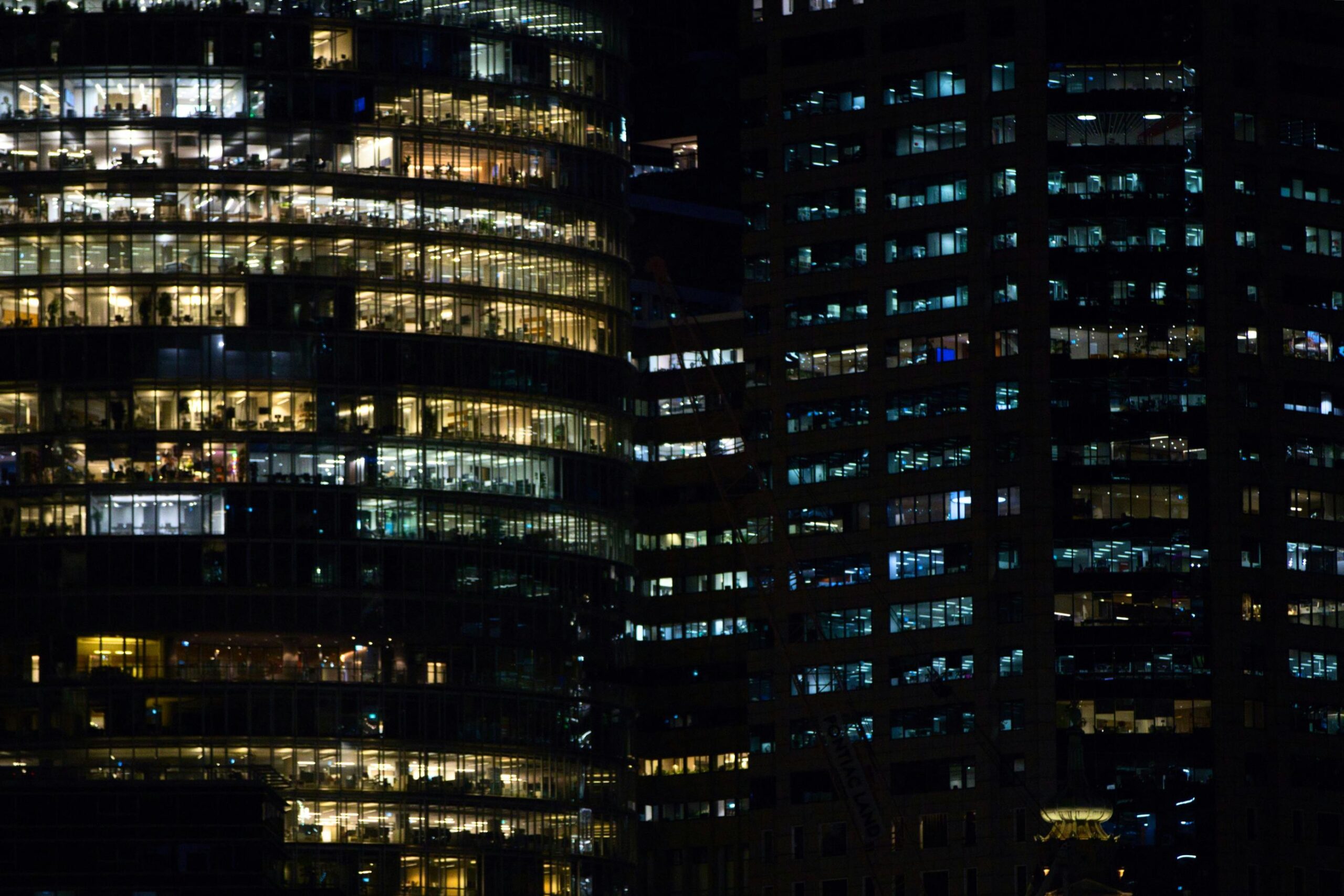 This image captures a dense night view of illuminated office buildings with glowing windows revealing busy interior workspaces. The contrasting warm yellow and cool blue lights create a rhythmic, geometric pattern against the dark city skyline.