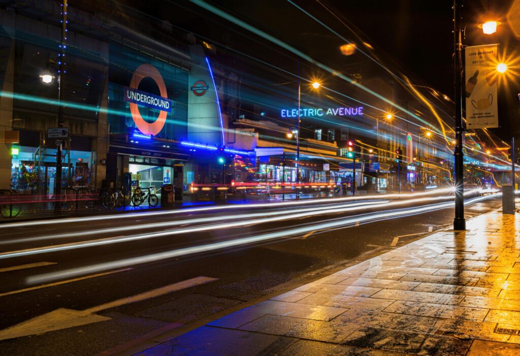 A long-exposure shot captures vibrant light trails from passing vehicles in front of Brixton Underground station at night. 