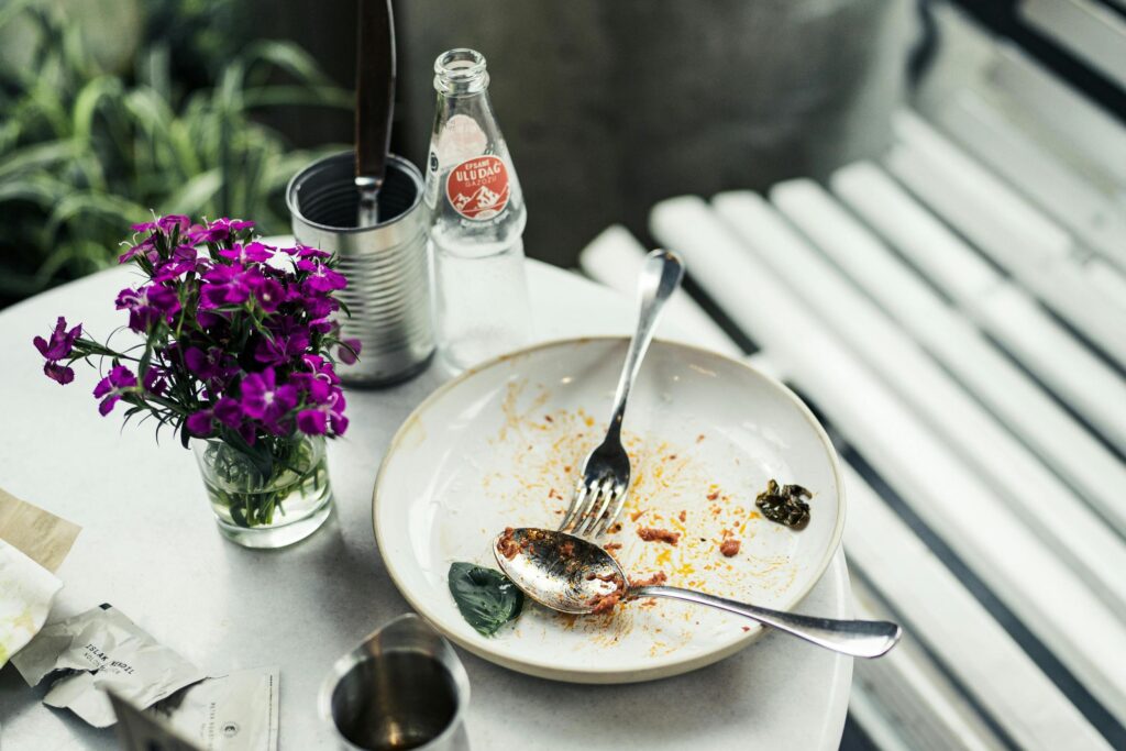 An empty, stained plate with a silver spoon and fork sits on a white table next to a vibrant bouquet of purple flowers. The scene includes a glass bottle of Uludağ Gazoz and a metal tin, all captured with a shallow depth of field against a blurred background.