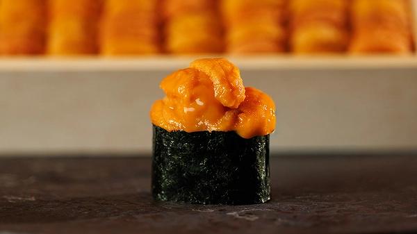 A single piece of uni gunkan sushi sits in the foreground, featuring bright orange sea urchin topped with a glossy glaze and wrapped in dark nori. In the blurred background, a wooden tray holds several additional rows of fresh sea urchin ready for preparation.