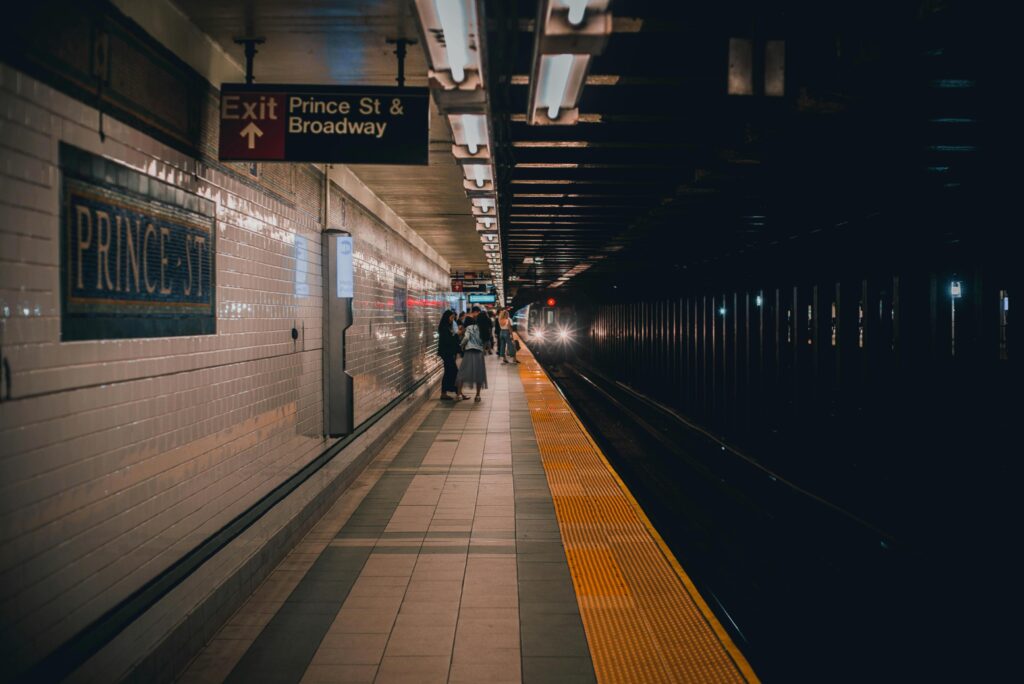 Commuters stand on a dimly lit subway platform at the Prince St & Broadway station as a train approaches in the distance. The scene captures the classic New York City underground atmosphere.