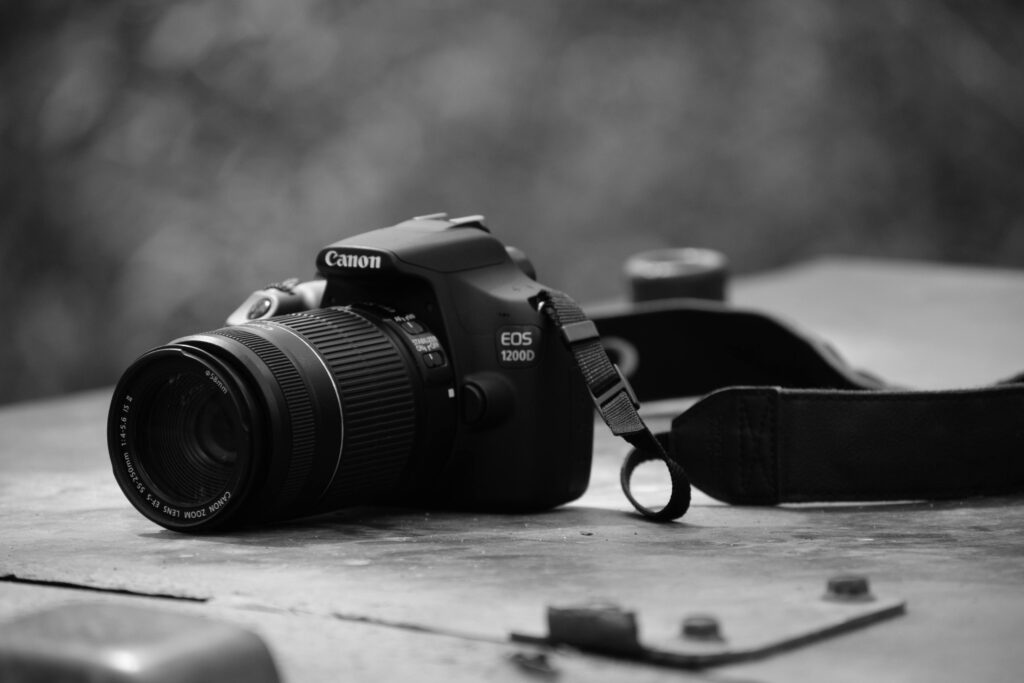 This black-and-white photograph features a Canon EOS 1200D DSLR camera equipped with a telephoto zoom lens. It sits on a weathered, flat surface with its neck strap draped to the side against a softly blurred background.
