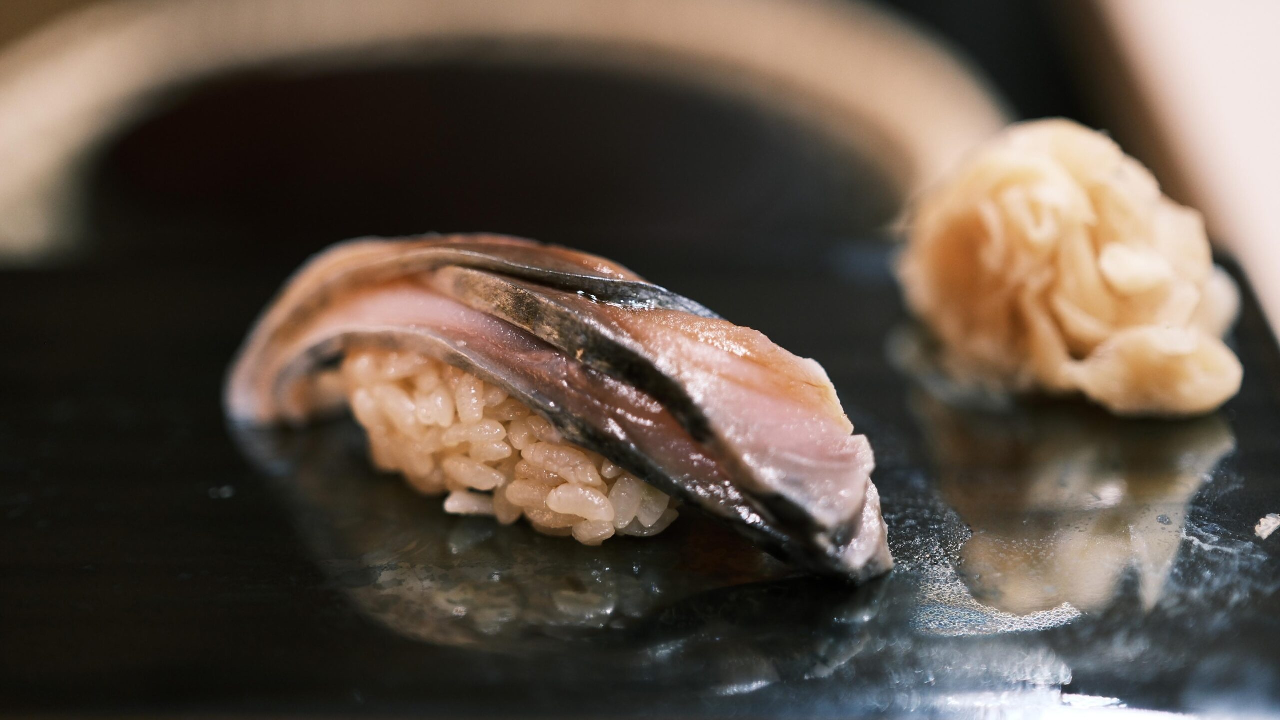 A single piece of saba nigiri rests on a sleek black platter alongside a delicate mound of pickled ginger. The fish features a silvery, cured skin and a deep pink hue, served over seasoned rice with a soft bokeh background.