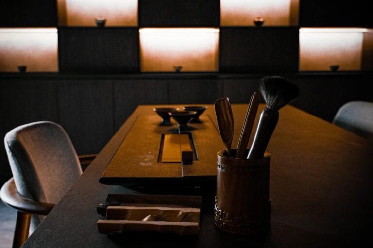 A collection of traditional tea tools and cups sits on a dark, minimalist table. Warm light glows from recessed shelving in the background, creating a moody and serene atmosphere.