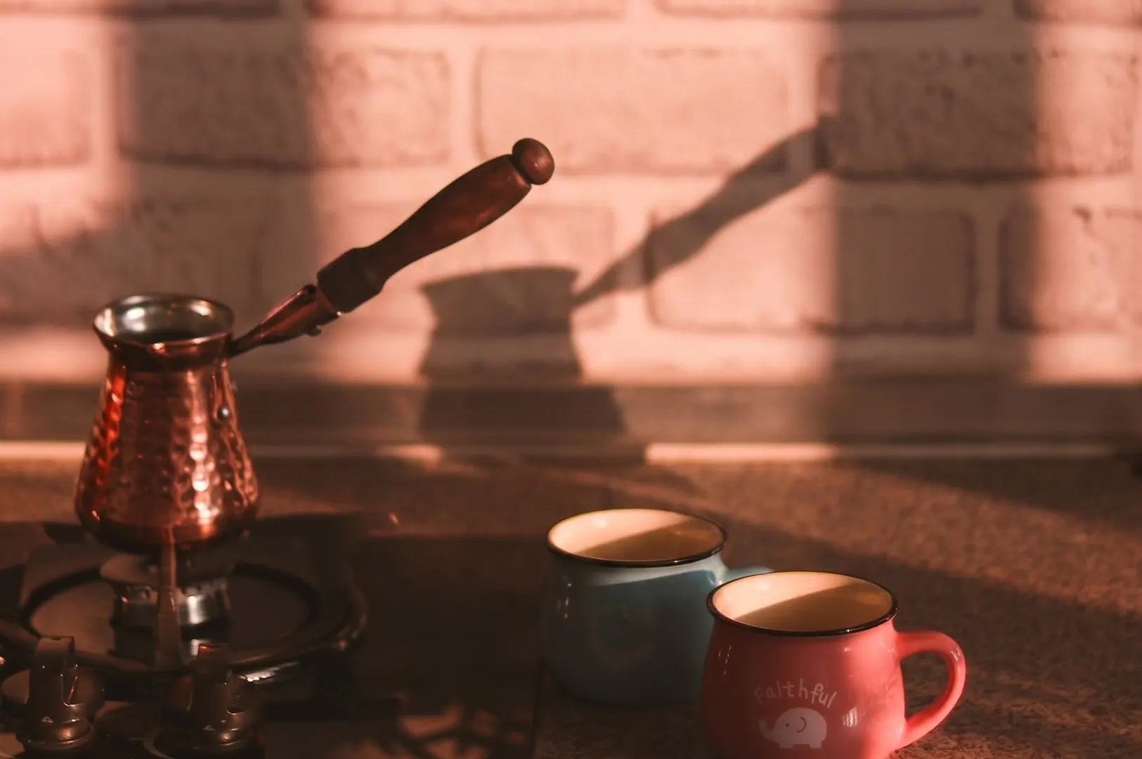Copper Turkish coffee pot on a stove with blue and pink mugs against a white brick wall in warm sunlight.