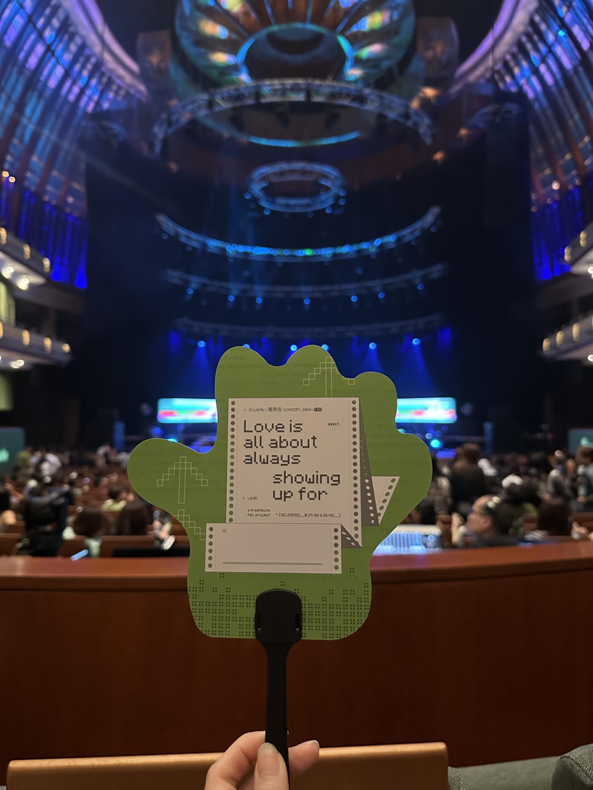 A hand holds up a green, hand-shaped fan featuring pixelated text that reads, "Love is all about always showing up for." The person is seated in a large, dimly lit concert hall with tiered seating and vibrant blue and purple stage lighting in the background.