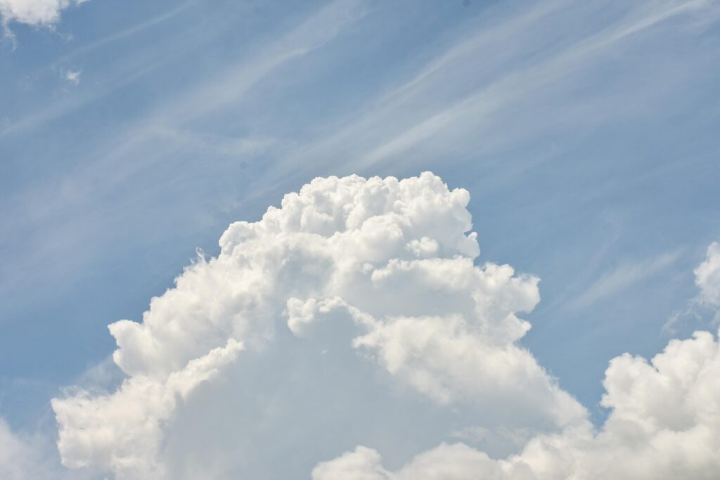 A massive, billowing white cumulus cloud dominates the center of the frame against a pale blue sky. Wispy, thin cirrus clouds streak diagonally across the background, adding texture to the bright atmosphere.