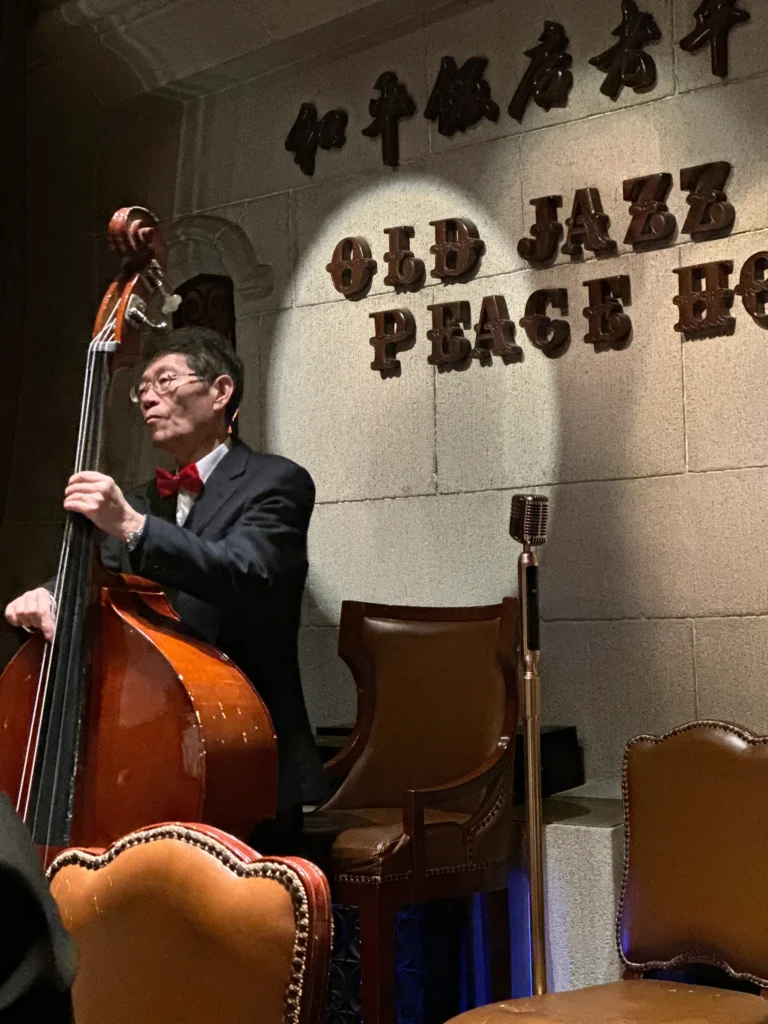 A double bass player in a tuxedo and red bowtie performs on a stage lit by a spotlight. The background features stone walls with signage for the Old Jazz Band at the Peace Hotel.