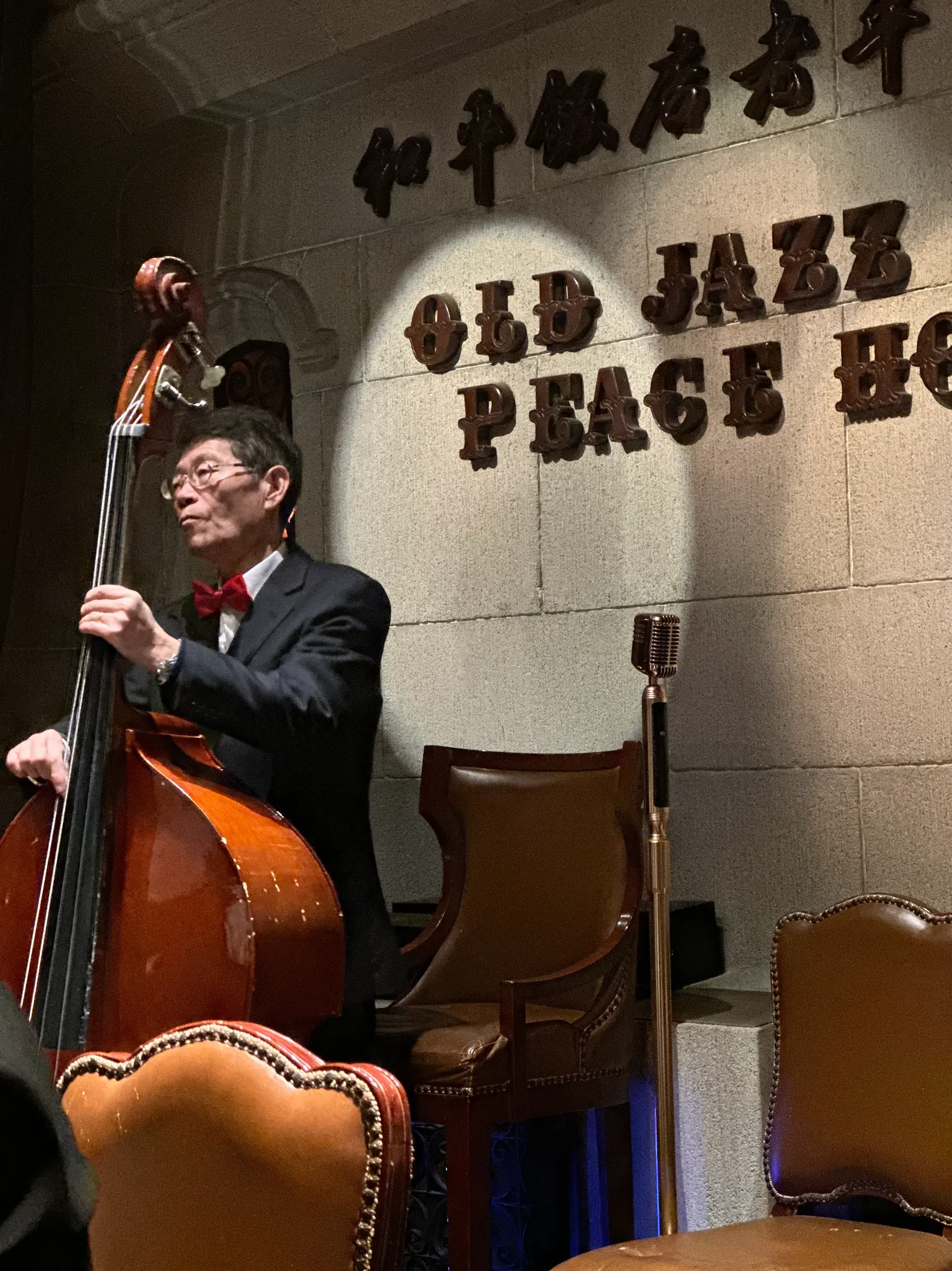 A double bass player in a tuxedo and red bowtie performs on a stage lit by a spotlight. The background features stone walls with signage for the Old Jazz Band at the Peace Hotel.