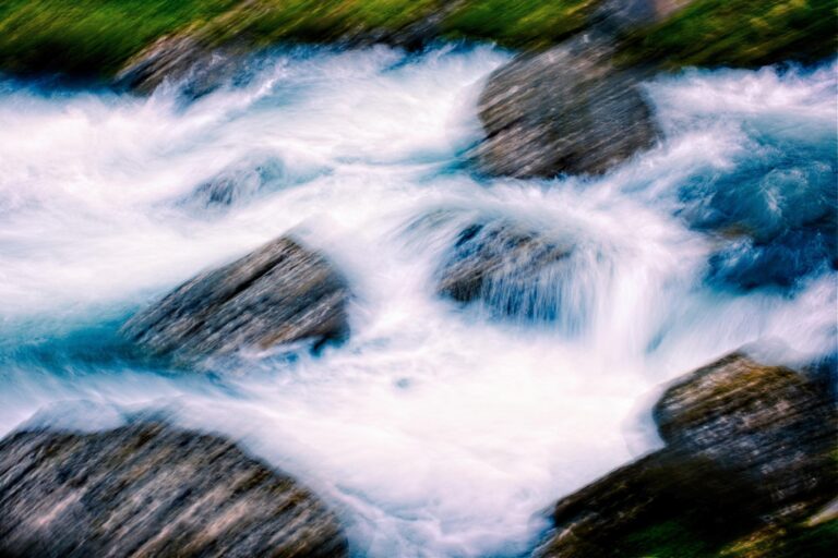 This high-angle shot captures turquoise water rushing over and around several large, grey rocks. The image features a strong motion blur that gives the frothing water a soft, ethereal texture and conveys a sense of rapid movement.