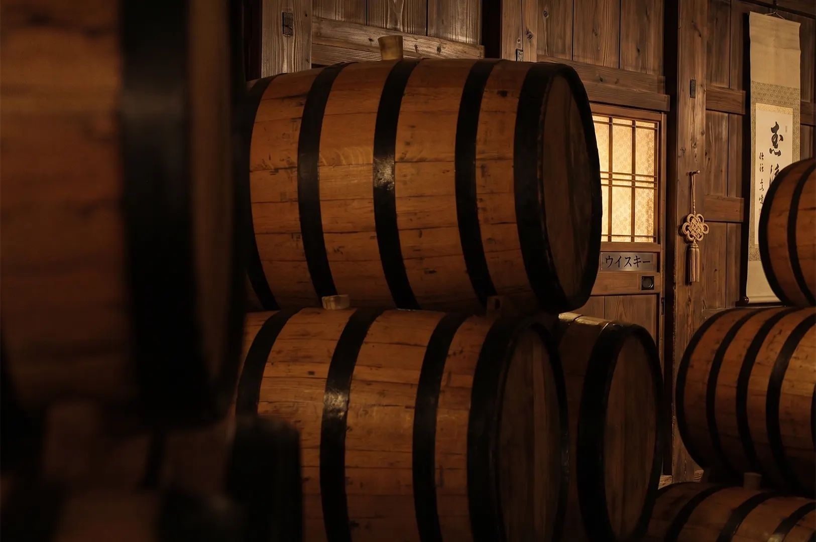 Stacked wooden whiskey barrels in a traditional Japanese distillery cellar with warm ambient lighting.