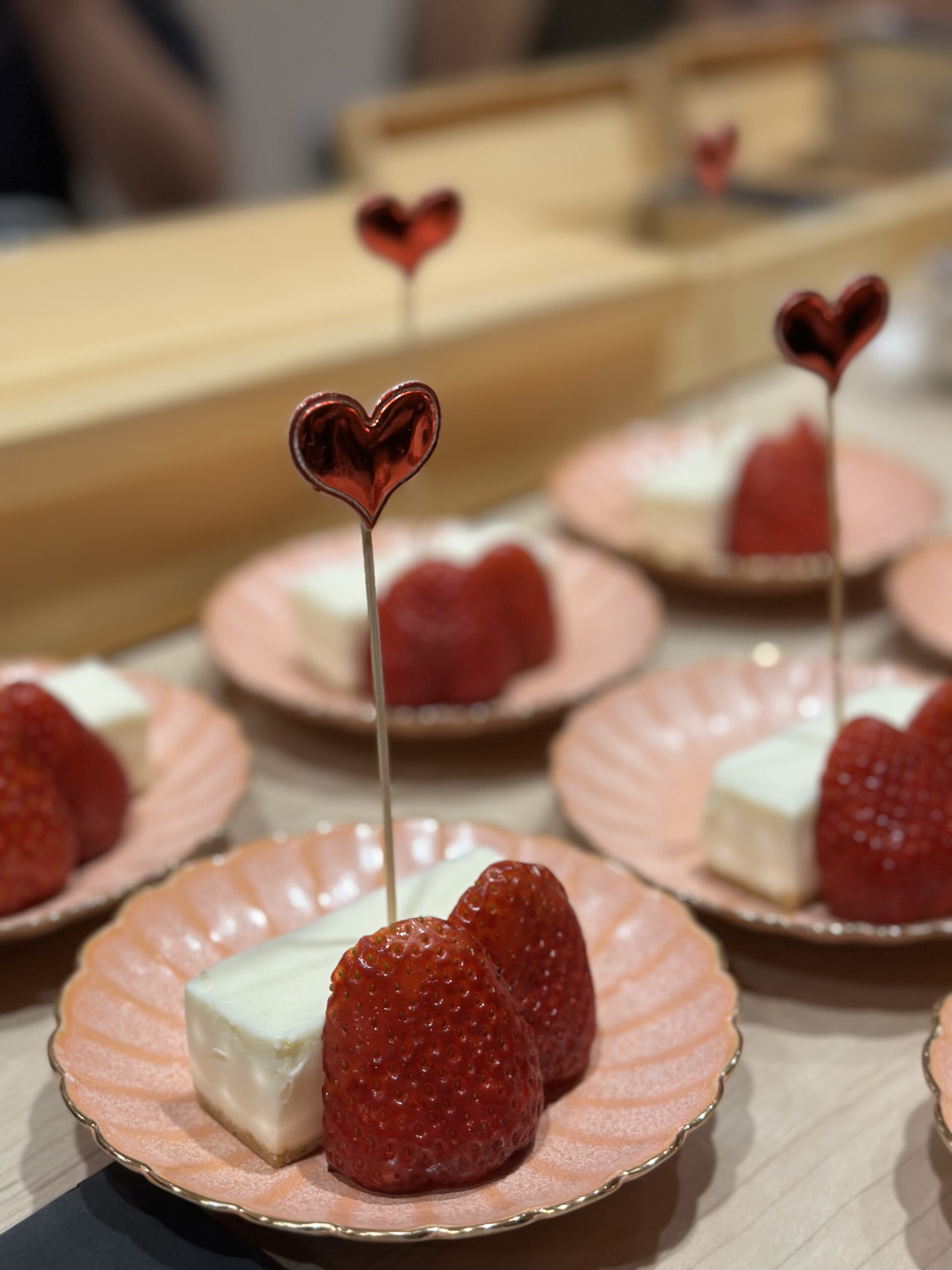 A close-up photograph of individual plates of strawberries and cheesecake bites with heart-shaped cocktail picks, arranged on a light wooden table.