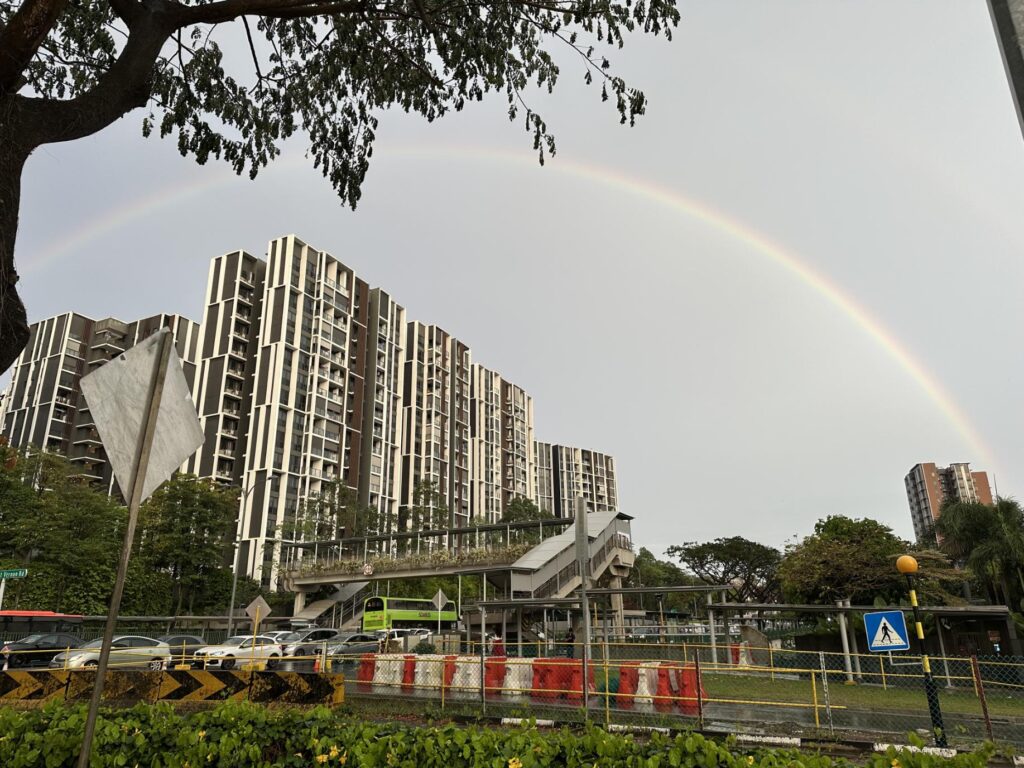 A vibrant rainbow arches across a gloomy, overcast sky, framing a modern multi-story residential complex and a nearby pedestrian bridge. In the foreground, a busy roadside features a green double-decker bus, parked cars, and temporary traffic barriers under the shade of large trees.