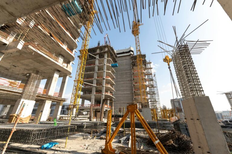 The image showcases a massive construction site featuring multiple high-rise buildings in various stages of skeletal development. Towering yellow cranes dominate the skyline above intricate frameworks of concrete, rebar, and scaffolding under a clear sky.