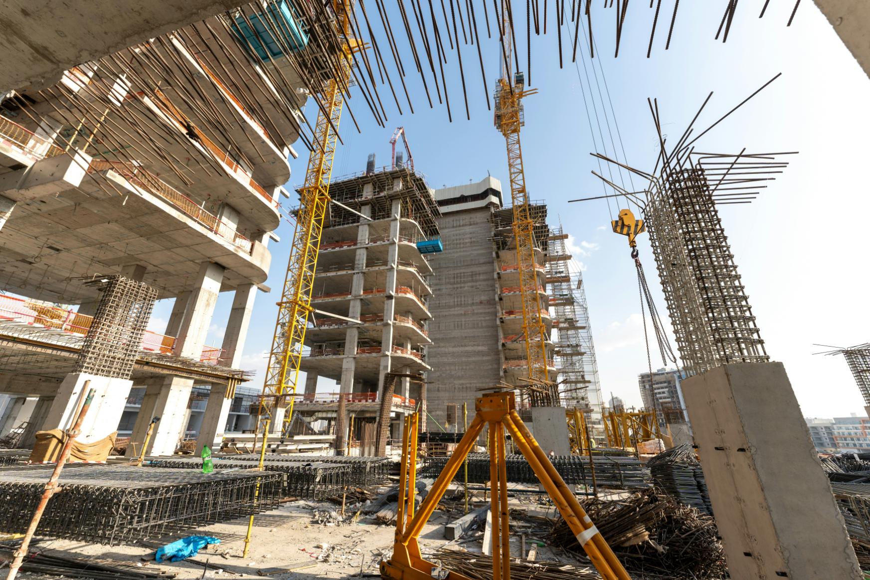 The image showcases a massive construction site featuring multiple high-rise buildings in various stages of skeletal development. Towering yellow cranes dominate the skyline above intricate frameworks of concrete, rebar, and scaffolding under a clear sky.