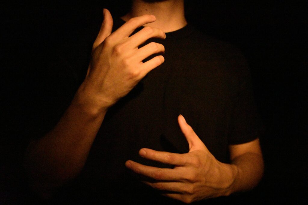 A person's hands are captured in a dramatic, low-light setting, illuminated by a warm orange glow against a dark background. One hand is raised near the chest while the other is positioned lower, creating an expressive and contemplative pose.