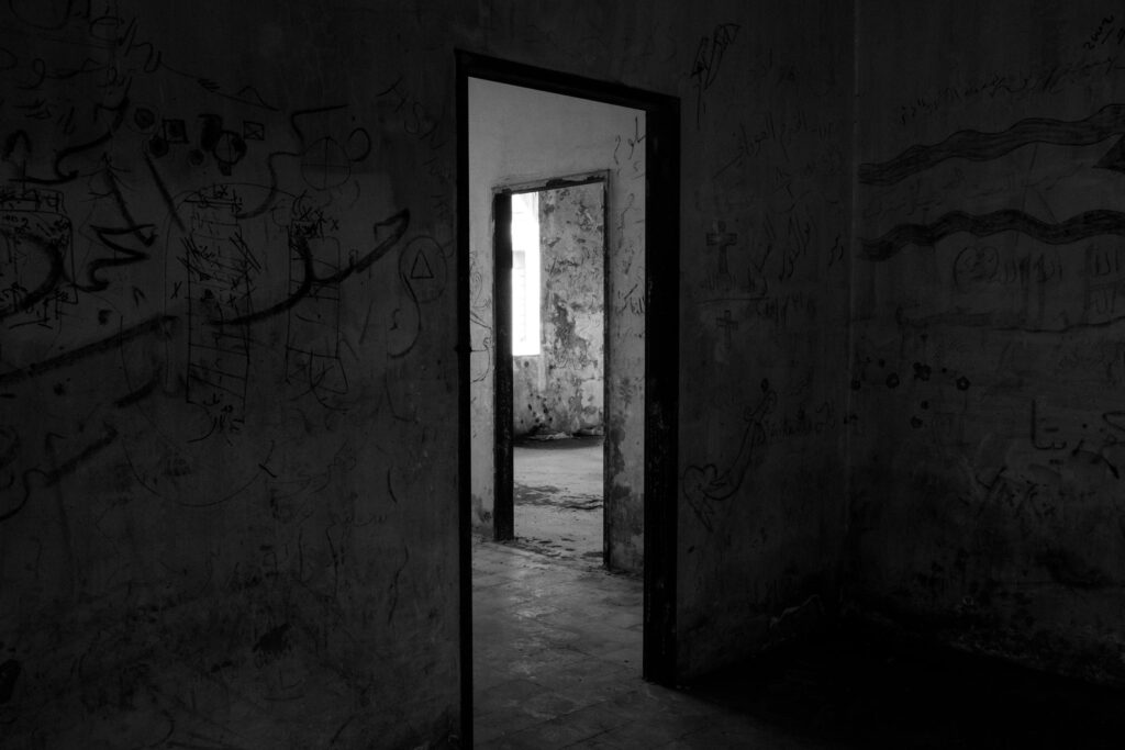 Graffiti-covered walls frame a dark room, leading the eye through a sequence of open doorways. A bright, distant window provides a stark contrast to the shadowy, abandoned interior.