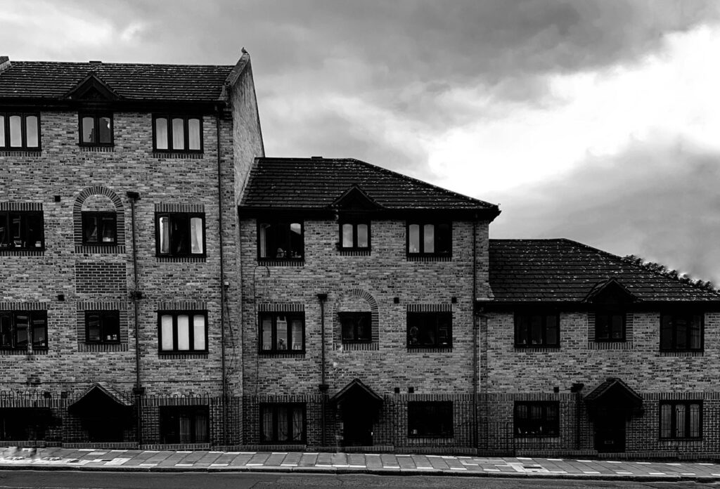 This black-and-white photograph showcases a multi-level brick residential building under a moody, overcast sky. The structure features a repeating pattern of dark-framed windows and gabled rooflines along a sloping sidewalk.