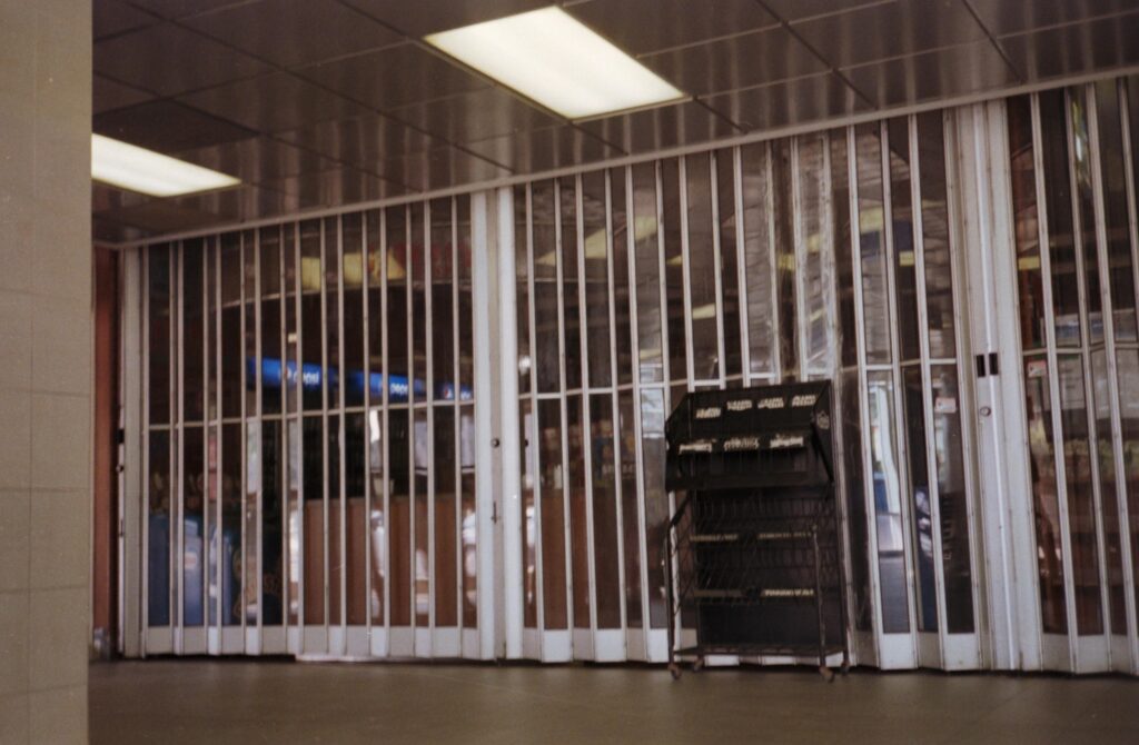 A storefront is closed behind a tall, translucent accordion gate under the glow of overhead fluorescent lights. In the foreground, an empty black wire display rack stands on a dark, polished floor next to a tiled pillar.