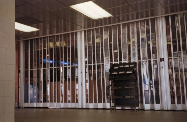 A storefront is closed behind a tall, translucent accordion gate under the glow of overhead fluorescent lights. In the foreground, an empty black wire display rack stands on a dark, polished floor next to a tiled pillar.