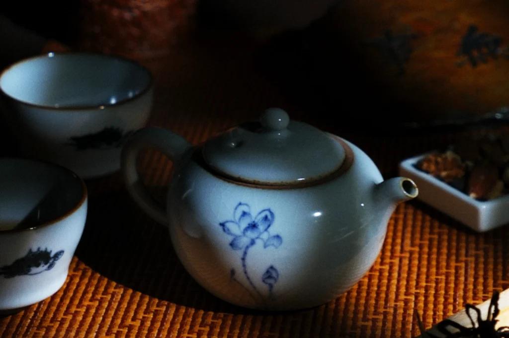 A ceramic teapot with a blue flower design sits on a woven mat alongside matching tea cups. Dramatic light highlights the textures of the teaware and the surrounding dark, cozy setting.