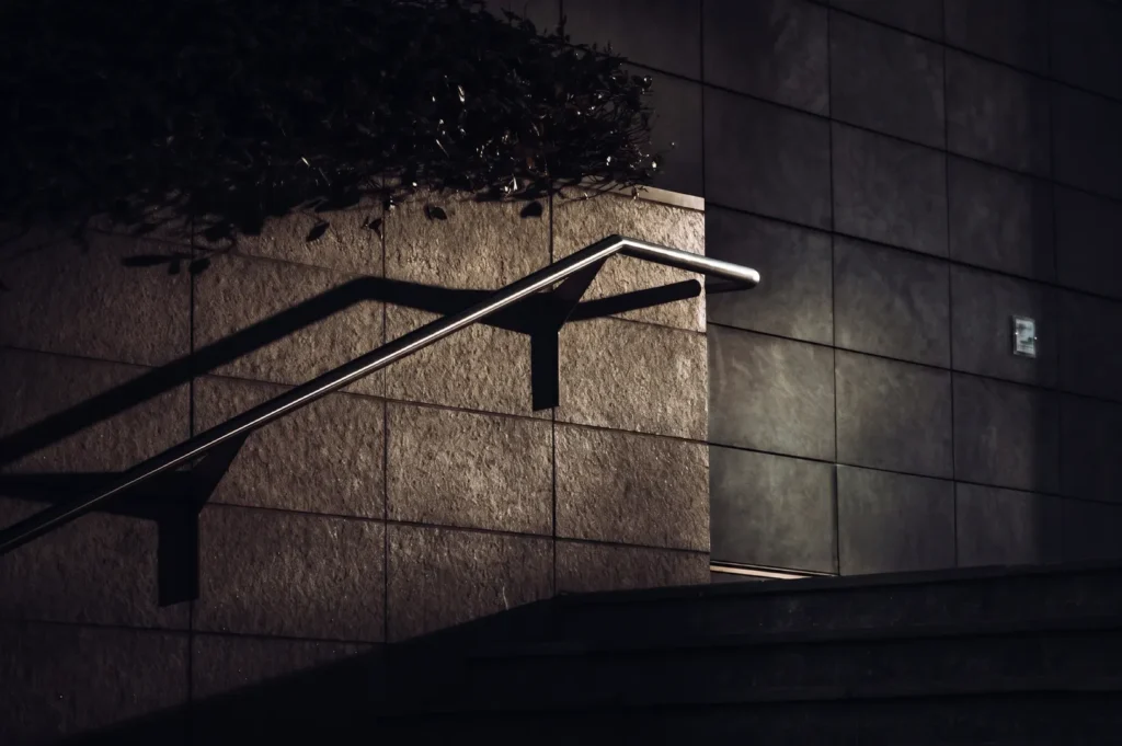 Modern metal handrail and stone stairs against a dark tiled building exterior with dramatic shadows.