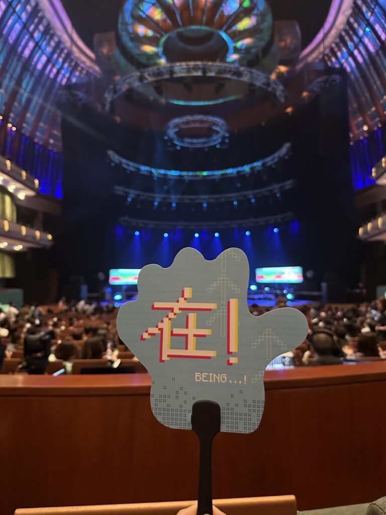 A light blue handheld fan shaped like a hand is held up in the foreground, featuring the Chinese character "在" and the English text "BEING...!" in a pixelated font. The background shows a large, crowded theater or concert hall with tiered seating and a stage illuminated by vibrant blue spotlights.