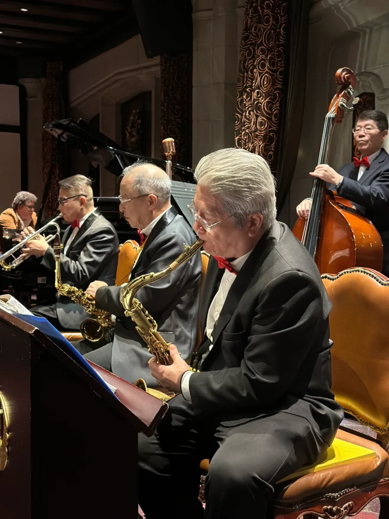 A formal jazz band performs in an elegant venue with musicians playing the saxophone, trumpet, and double bass. The performers are dressed in matching black suits and red bowties while seated in ornate, yellow-cushioned chairs.