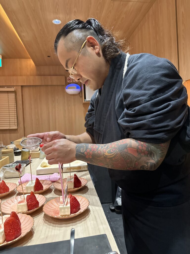 A chef with a floral tattoo sleeve dusting powdered sugar over strawberry and cheesecake desserts.