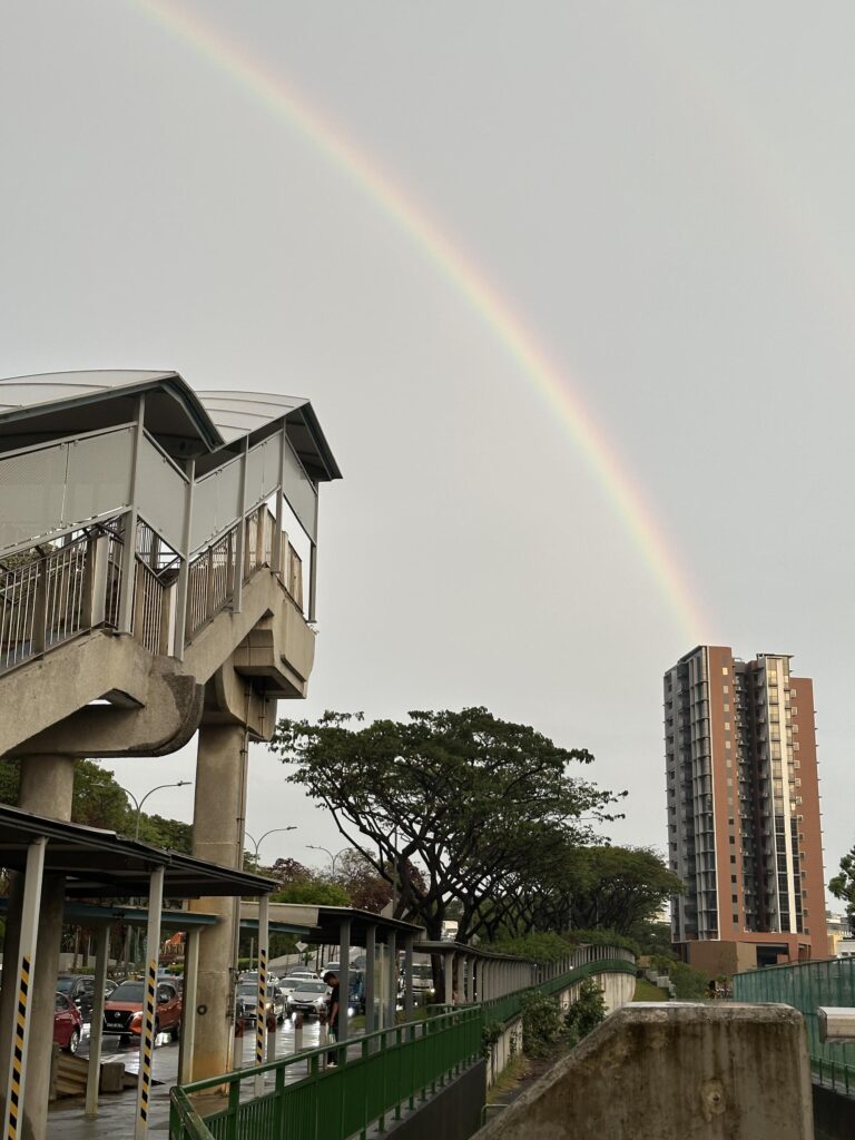 A bright, vivid rainbow arches across a gray, overcast sky, appearing to end behind a tall modern apartment building. In the foreground, a concrete pedestrian overpass and a sheltered walkway line a wet street filled with cars.