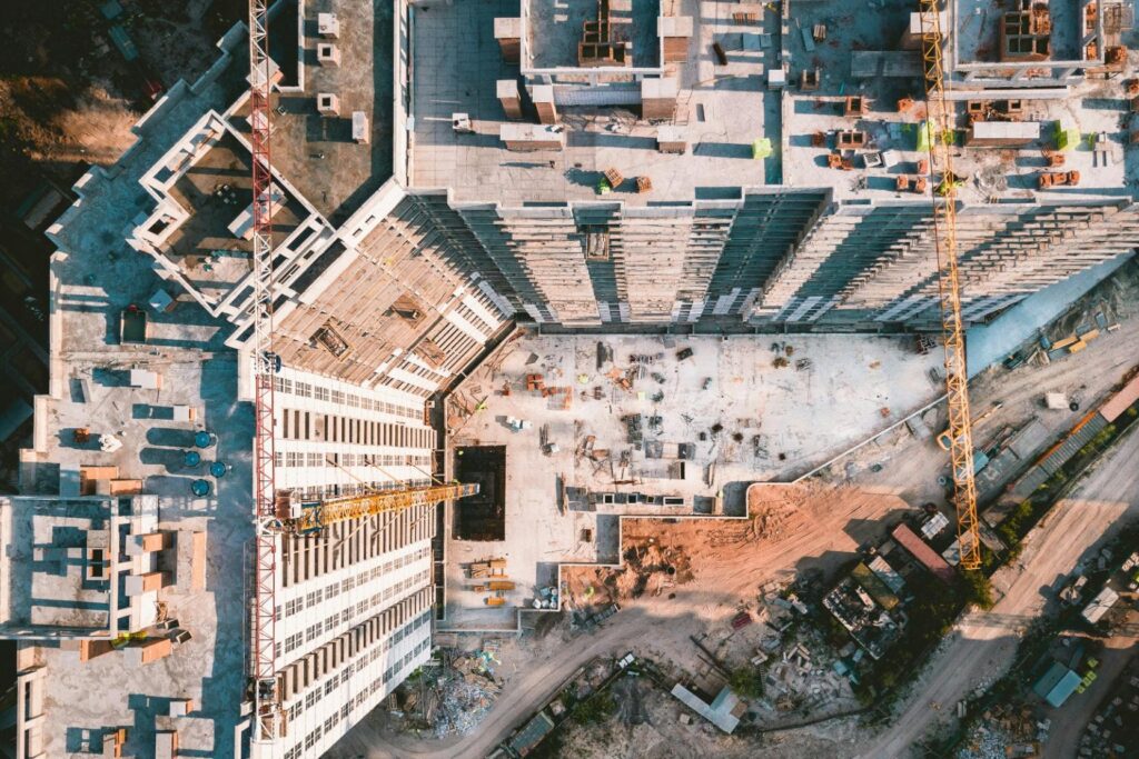 This aerial photograph captures a high-rise construction site from a top-down perspective, highlighting the geometric patterns of the building's concrete framework. Long shadows from the towering yellow cranes stretch across the dirt and scaffolding.