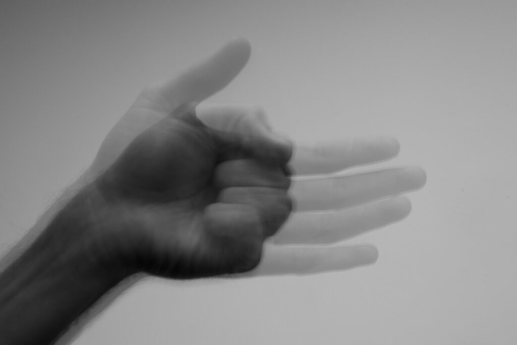 This black and white photograph captures a hand in motion, creating a ghostly, blurred trail as it transitions from an open palm to a clenched fist. The overlapping, translucent layers against the minimalist background.
