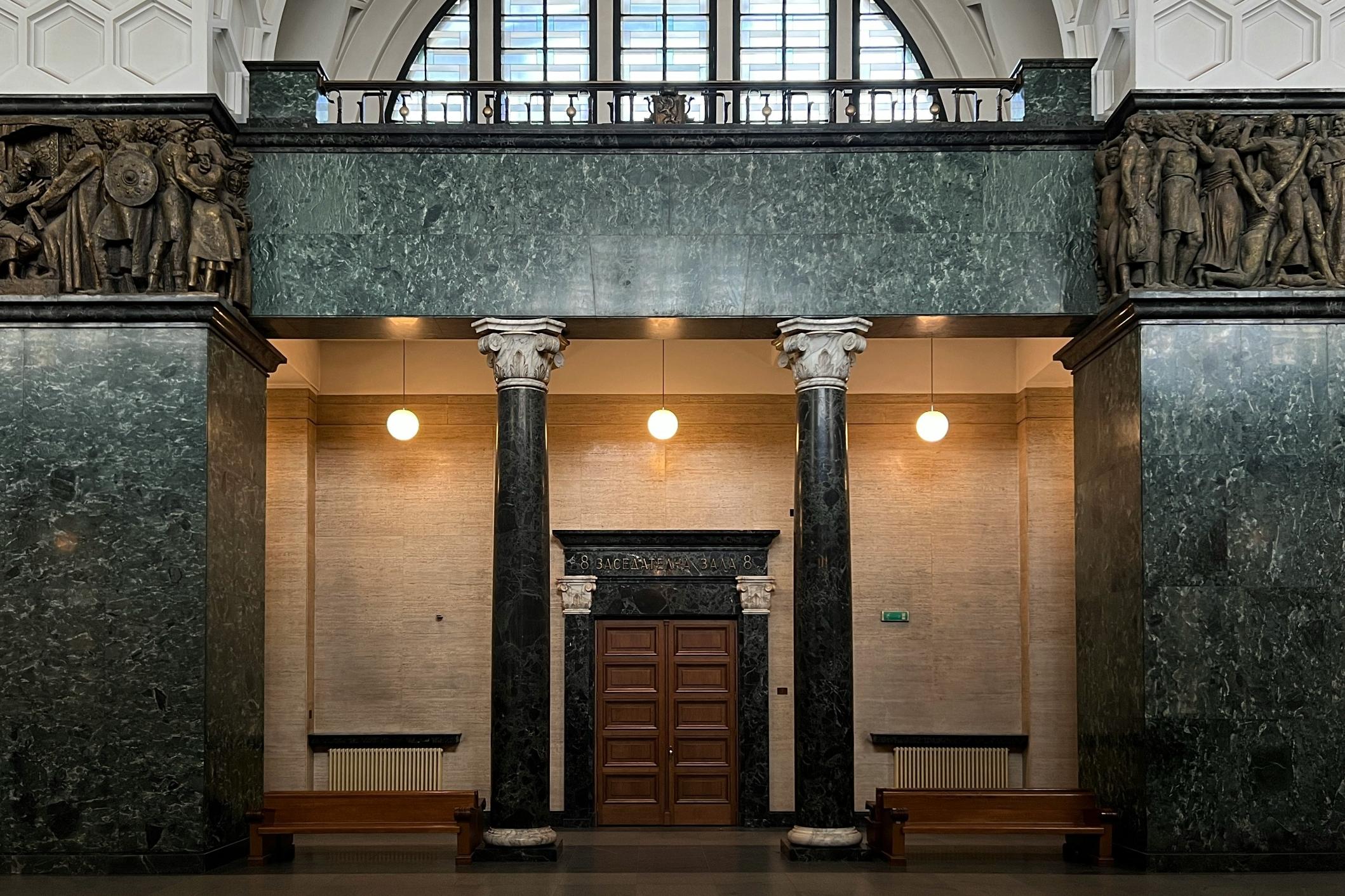 Grand marble pillars and intricate friezes frame the entrance to a stately hall. Warm pendant lights glow above a central wooden door, casting a soft light across the stone interior.