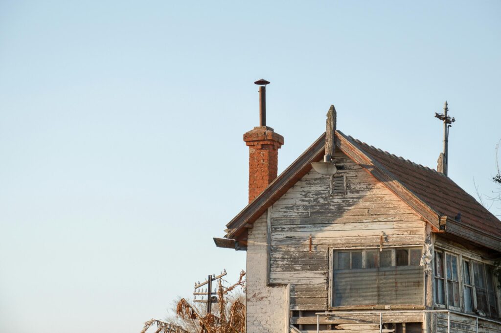 An weathered, two-story building with peeling white paint stands against a clear, pale blue sky. A prominent brick chimney rises from the gabled roof, which is flanked by an old-fashioned utility pole and several perched birds.