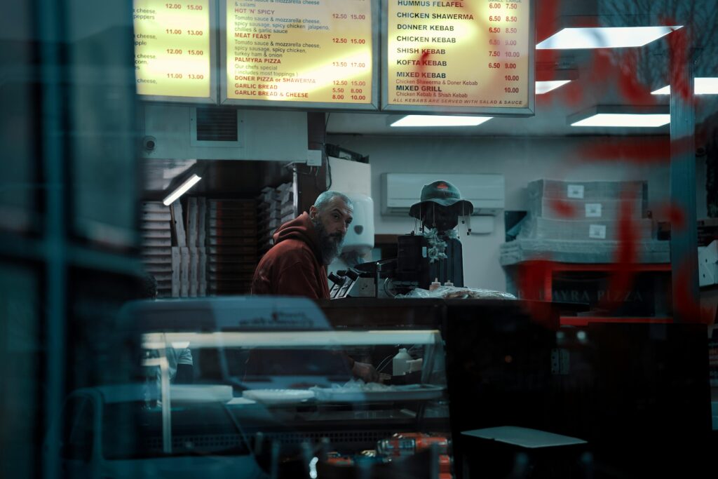 A man with a beard and a red hoodie stands behind the counter of a dimly lit kebab shop, glancing toward the window. The scene is captured from outside through glass reflecting red graffiti, showing glowing menu boards and stacked pizza boxes in the background.